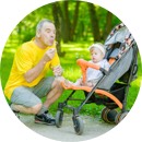 imagen de un abuelo soplando una flor de diente de león a lado de su nieta, quien es una bébé que está sentada en un coche de ruedas